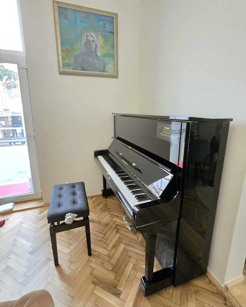 Fekete Yamaha pianínó és egy zongorapad egy halszálkás parkettás szobában